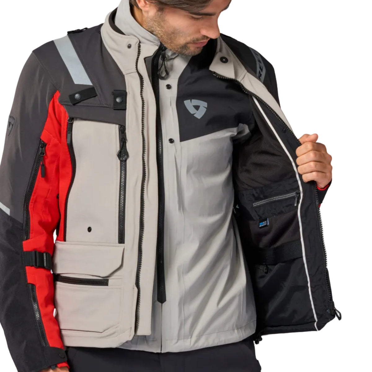A man wearing a technical outdoor jacket, opening the front zipper to show the black interior lining. The jacket features a light gray body, black sleeves and collar, and red side panels, with a small shield logo on the left chest.