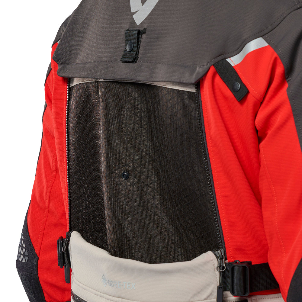 Close-up of the back of a technical outdoor jacket in red, dark gray, black, and beige, showing a mesh back panel, buckle closure, and a front zip pouch.
