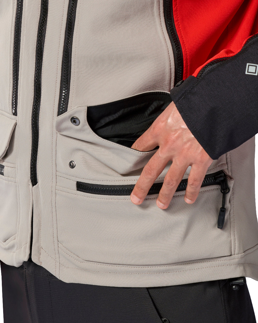 Close-up of a beige technical jacket with a red inner panel, black zippers, and a hand opening a front zip pocket.