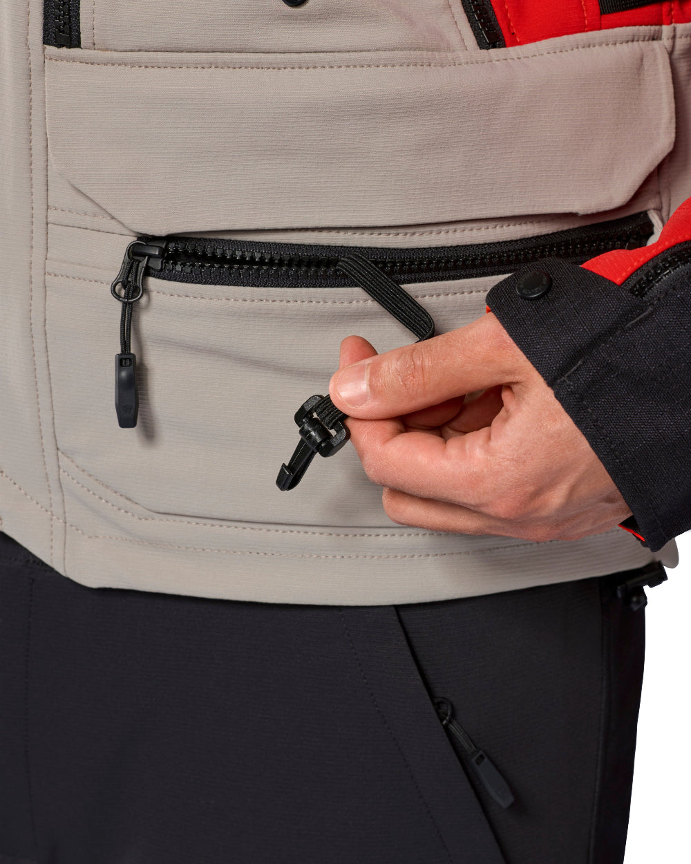 Close-up of a person wearing a light beige technical jacket with a black zipper pocket and a hand holding a black zipper pull tab, with dark pants and a red jacket lining visible.