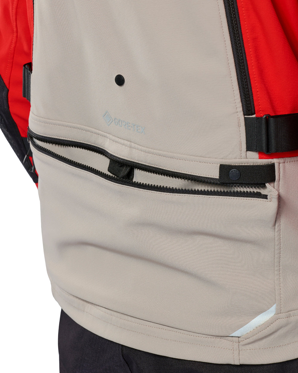 Close-up of a beige GORE-TEX jacket panel with a black zipper pocket, black buckle strap, and a red outer layer visible on the left side.
