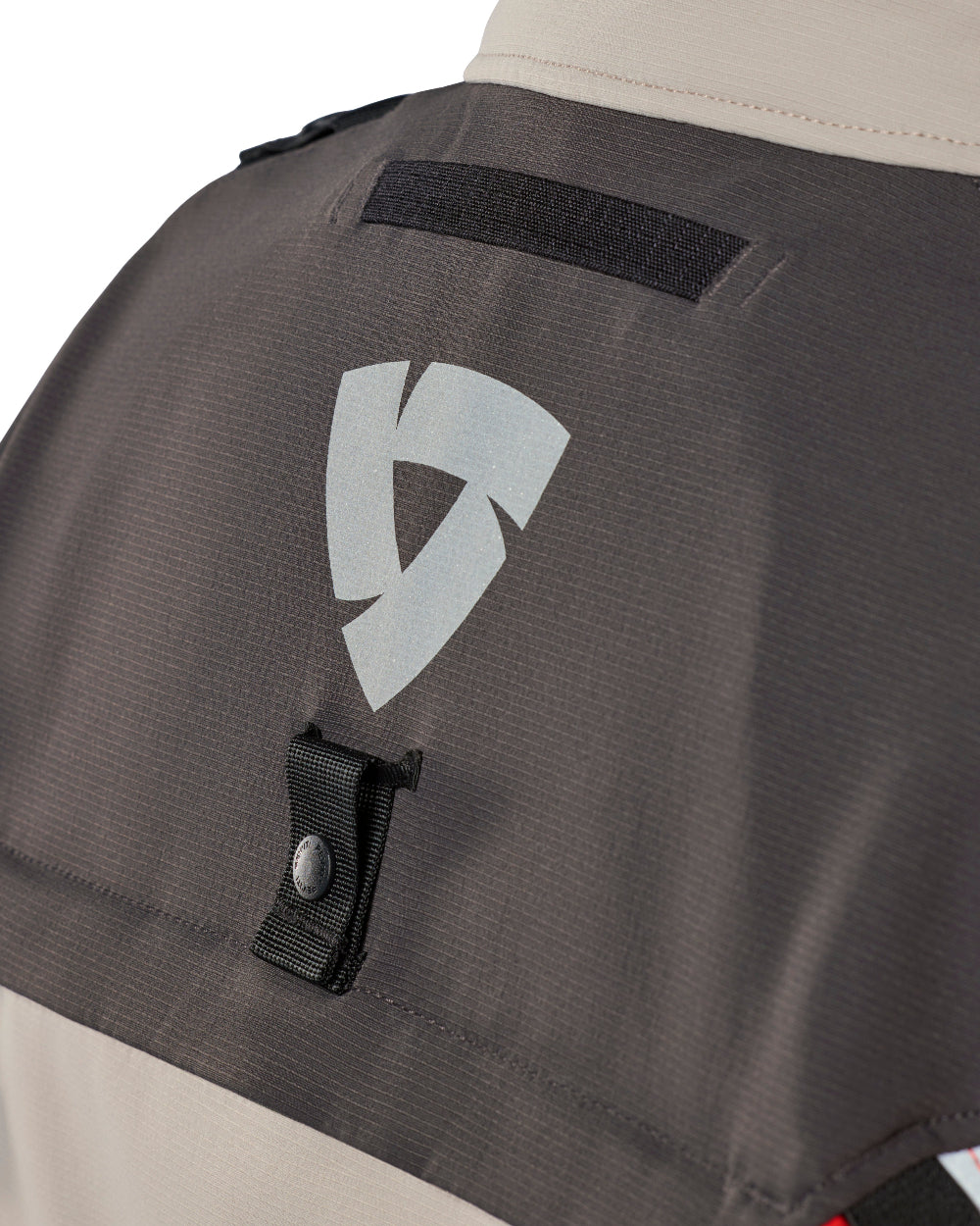 Close-up of a dark gray technical jacket or vest showing the back panel with a reflective silver geometric logo, a black hook-and-loop strap, and a light beige collar and lower section.