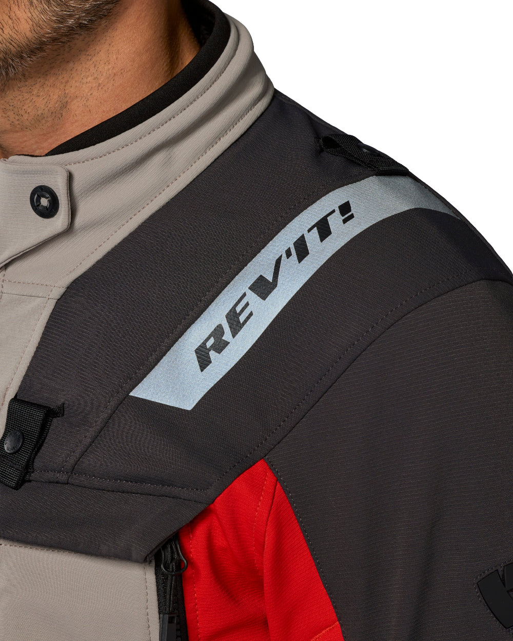 Close-up of a motorcycle jacket showing the shoulder area with a light gray and black REV'IT! logo on a dark gray panel, a beige collar, and a red lower panel.
