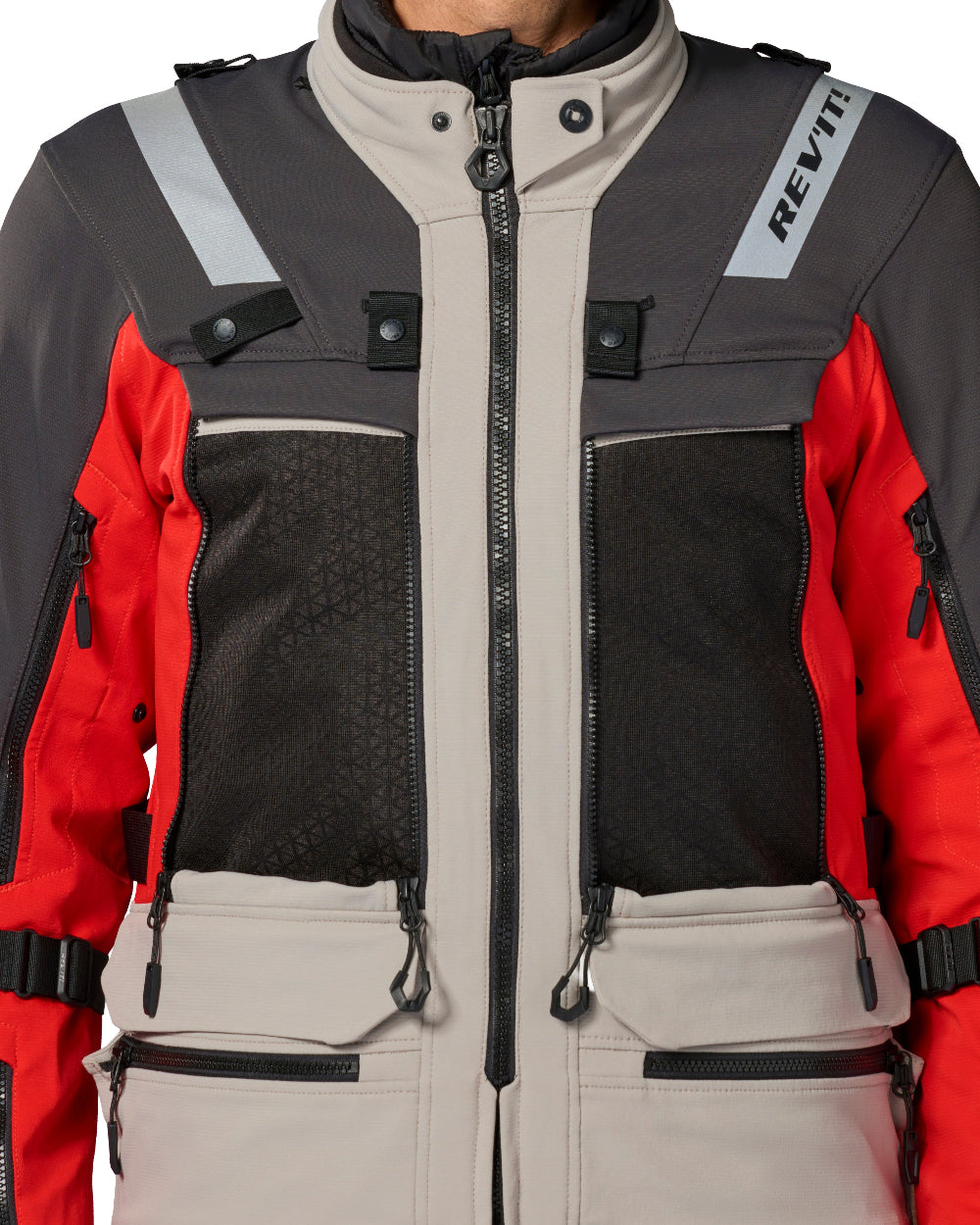Close-up of a technical riding jacket with a color-blocked design in beige, dark gray, and red. Features a full-length front zipper, multiple zippered pockets, reflective gray shoulder stripes, and the REV'IT! brand name printed on a white sleeve patch.