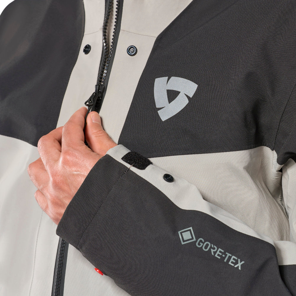 Close-up of a person's hand adjusting the zipper on a colorblock jacket, showing a dark gray and off-white design with a visible GORE-TEX logo on the sleeve.