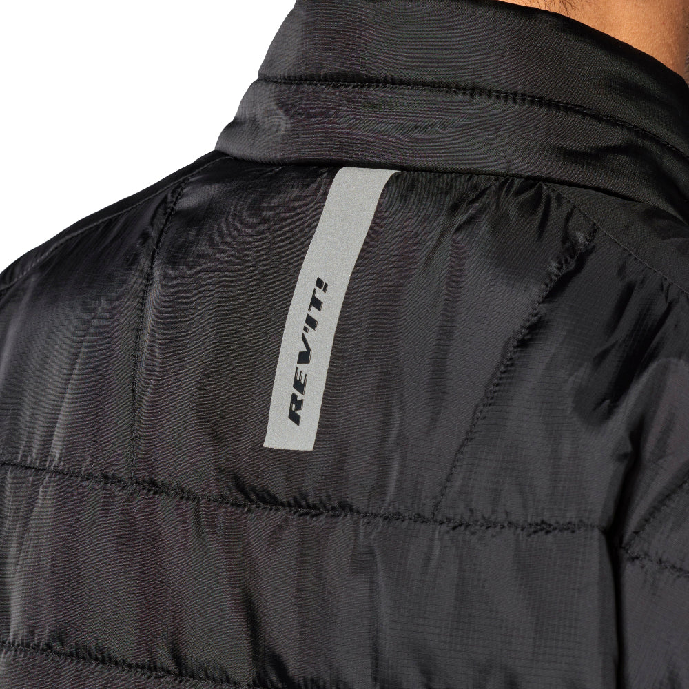 Close-up of the back collar and upper back of a black quilted puffer jacket, featuring a vertical light gray reflective strip with the brand name REV'IT! printed vertically in black.