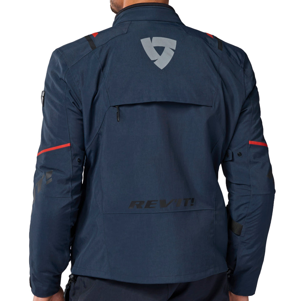 A man viewed from the back wearing a dark navy technical jacket with a gray shield logo on the upper back and red accent lines on the sleeves, paired with matching dark pants, against a plain white background.