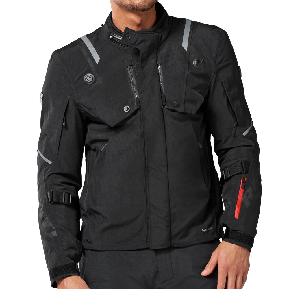 A man wearing a black technical motorcycle jacket and matching black pants, photographed against a plain white studio background.
