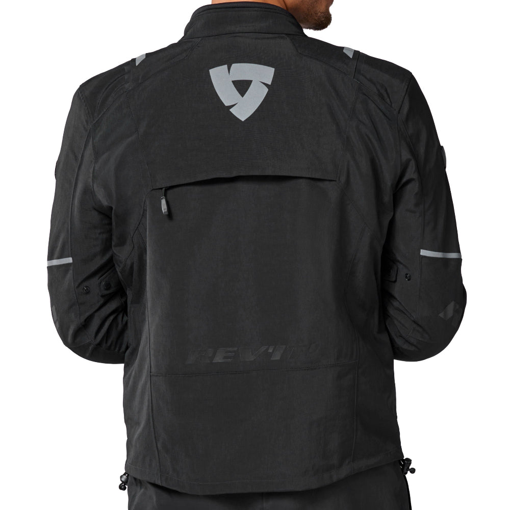 A man viewed from the back wearing a black motorcycle jacket with a gray shield logo on the upper back, paired with matching black pants, against a plain white background.