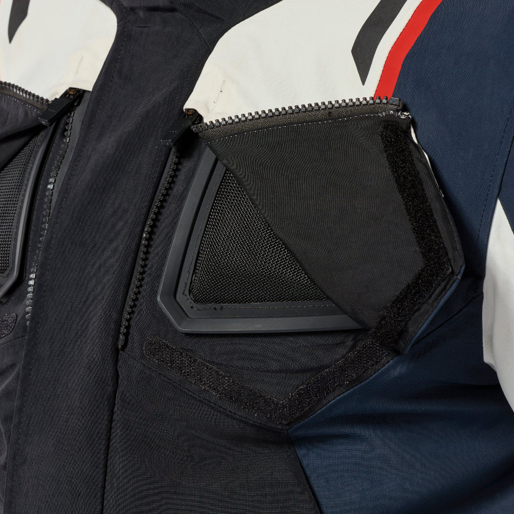 Close-up detail of a technical motorcycle jacket showing a black zippered chest pocket with a mesh insert and hook-and-loop flap, alongside navy, white, and red color-blocked fabric panels.