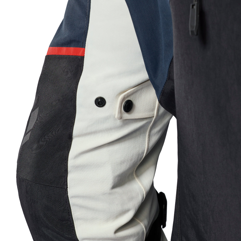 Close-up of a color-blocked technical jacket or pants panel, showing navy blue, off-white, and dark gray sections with a small red stripe and snap-button flap detail.