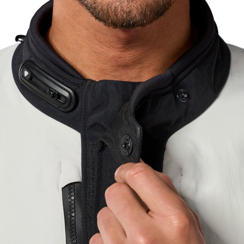Close-up of a man wearing a black and white jacket, adjusting the collar with his hand. The jacket features a high black collar with snap buttons and a black zipper.