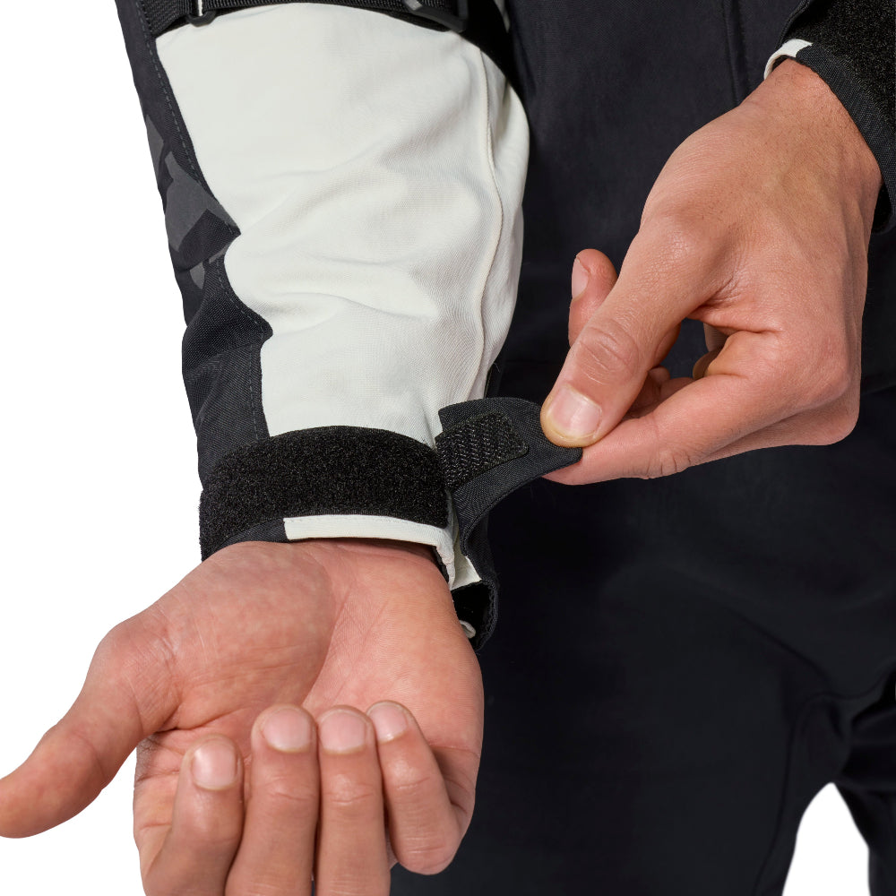 Close-up of a person wearing a black wrist strap and velcro cuff on a white and black sleeve, with the other hand adjusting the strap.