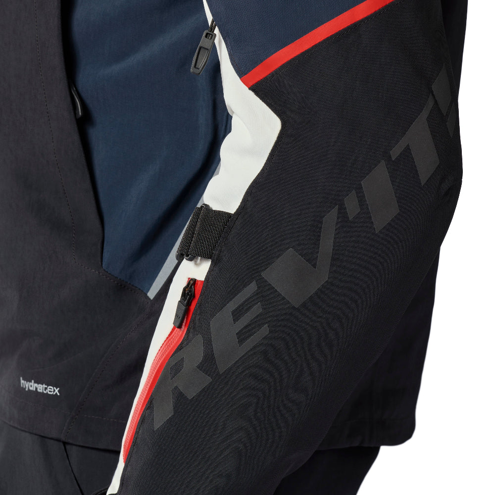 Close-up of a technical jacket sleeve showing navy, black, white, and red color blocking with a large dark gray REV'IT! logo on the forearm.