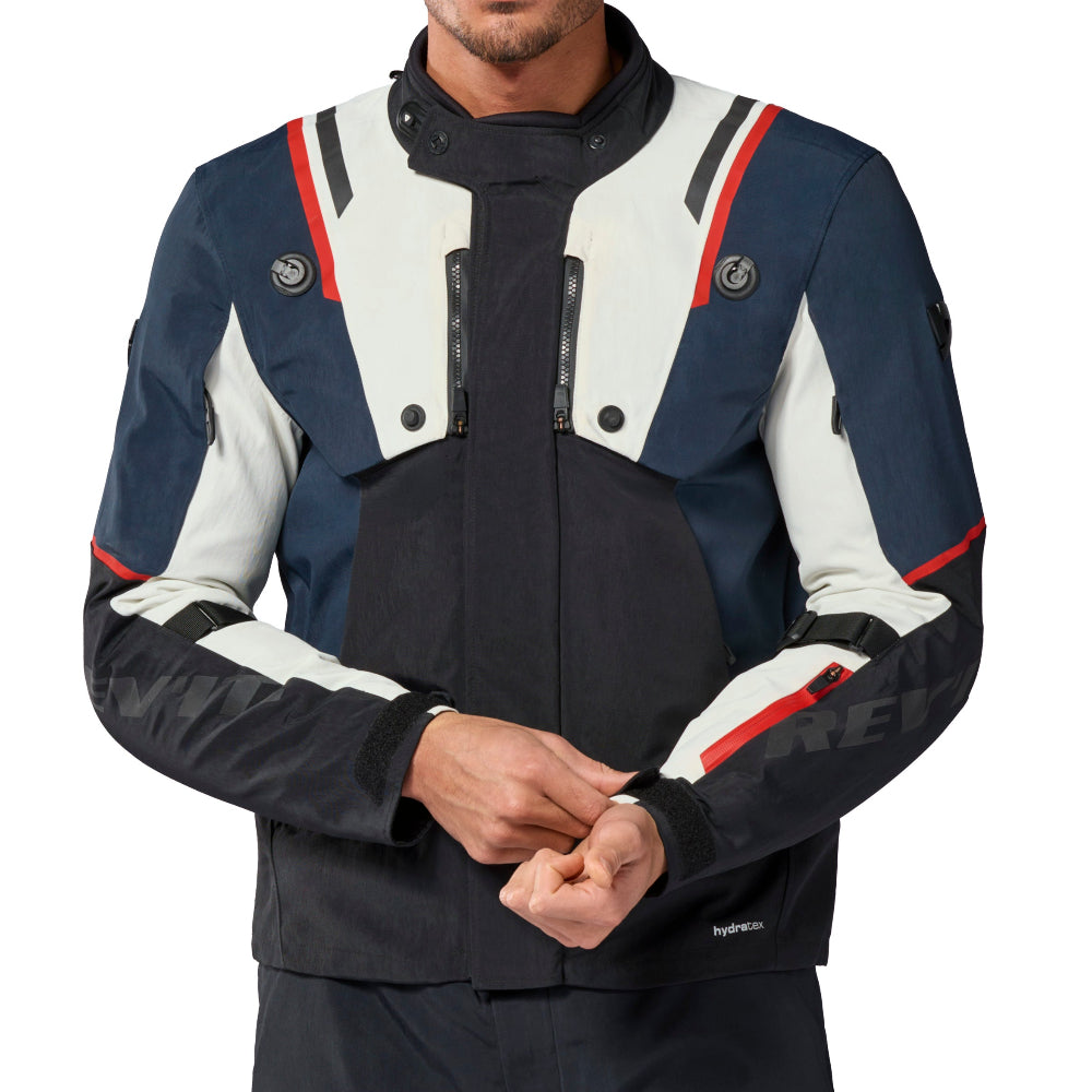 A man modeling a technical motorcycle jacket and matching pants against a plain white background. The jacket features a color-blocked design in navy blue, white, and black with red and white stripe accents on the shoulders and sleeves.