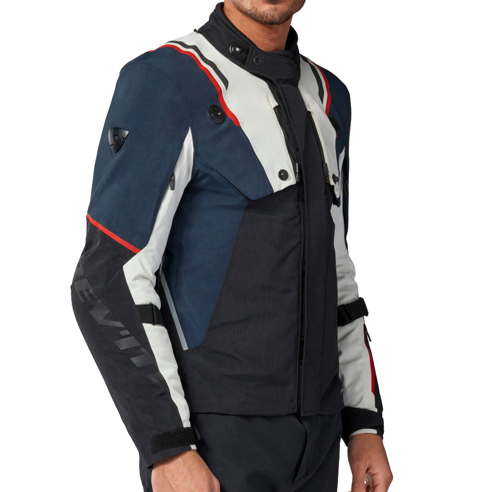 A man modeling a color-blocked motorcycle jacket with navy blue, white, and black panels, accented with thin red stripes, against a plain white background.