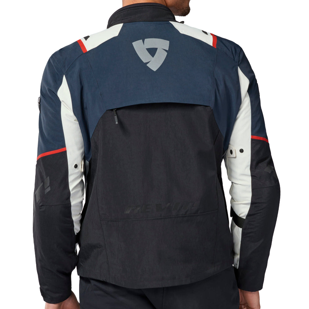 A man viewed from the back wearing a technical motorcycle jacket with navy, white, and red panels, featuring a large shield logo on the upper back.