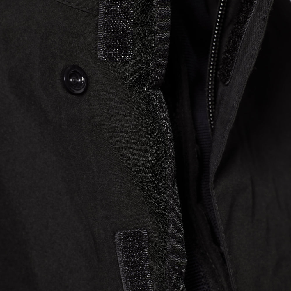 Close-up detail of a black jacket showing a button closure, zipper track, and textured hook-and-loop fastening panels.
