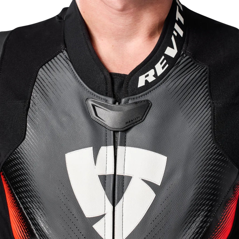Close-up of a person wearing a black and dark gray motorcycle leather jacket with a white REV'IT! logo on the collar and a large white shield logo on the chest.