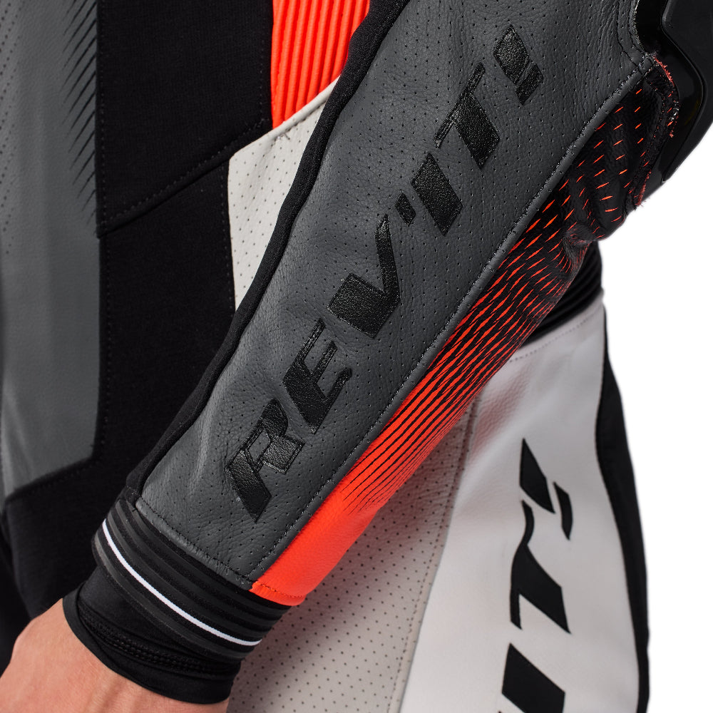 Close-up of a motorcycle racing suit sleeve with a dark gray leather panel, black REV'IT! branding, and bright orange-red accents, worn by a rider.