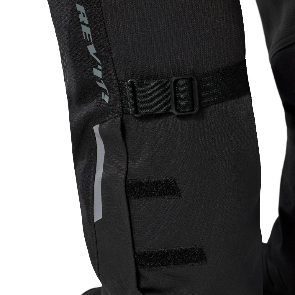 Close-up of a black technical riding garment leg with a buckle strap, hook-and-loop fasteners, and a vertical gray REVT logo on a white background.