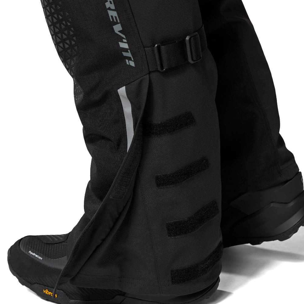 Black REV'IT! outdoor boot cover with hook-and-loop straps and a buckle closure, shown on a person's lower leg with a black boot underneath, on a white background.