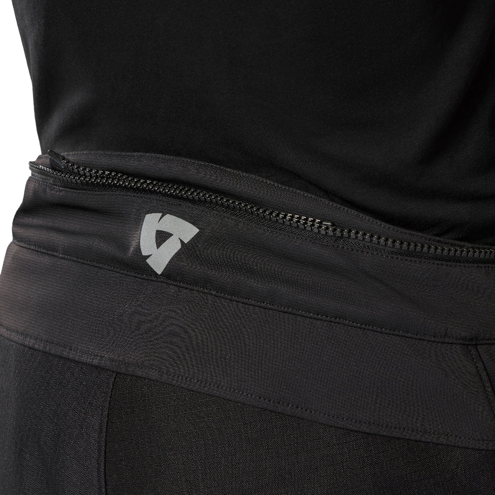 Close-up of a black athletic waistband with a horizontal jacket connection zippers and a small white geometric logo, shown on a person wearing black activewear.
