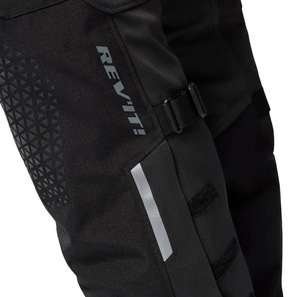 Close-up of black REV'IT! motorcycle pants showing the side panel with a gray reflective vertical stripe, a buckle strap, and textured geometric grip panels.