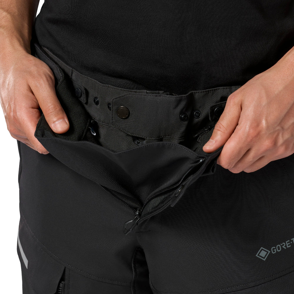 Close-up of a person wearing black technical pants and pulling open a waist zipper with both hands. A small Gore-Tex logo is visible near the lower right leg.