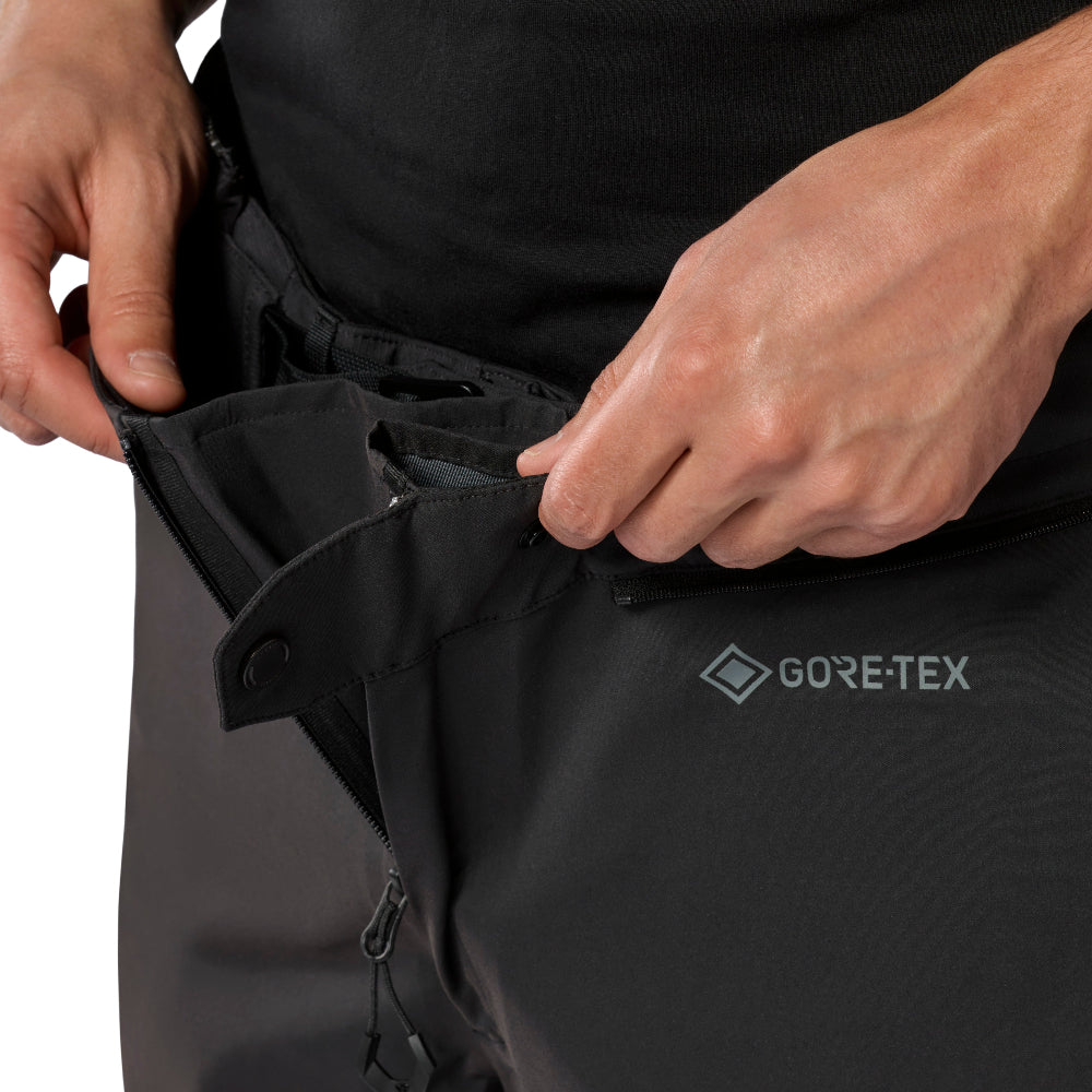 Close-up of a person's hands opening the waistband of black GORE-TEX pants, showing the adjustable hook-and-loop closure and the GORE-TEX logo on the side.