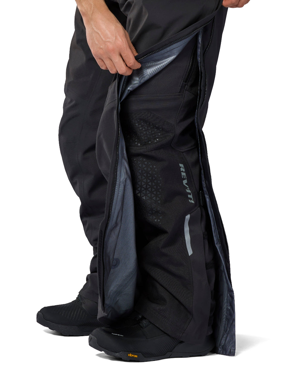 Close-up of a person wearing black REVIT waterproof liner over riding pants, lifting the left leg to show the interior lining and reflective branding on the lower leg.