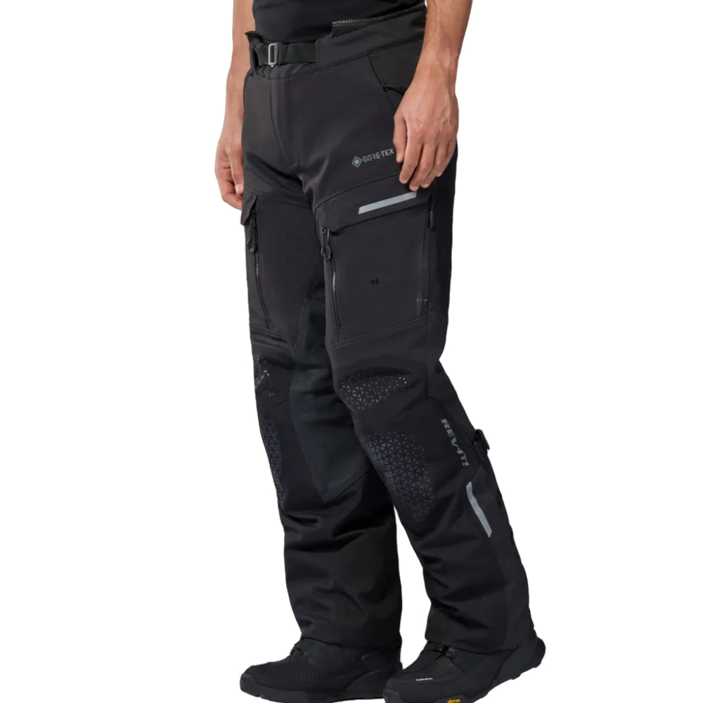 A person wearing black Gore-Tex outdoor pants with reflective gray zipper details and textured knee panels, shown from the waist down on a white studio background.