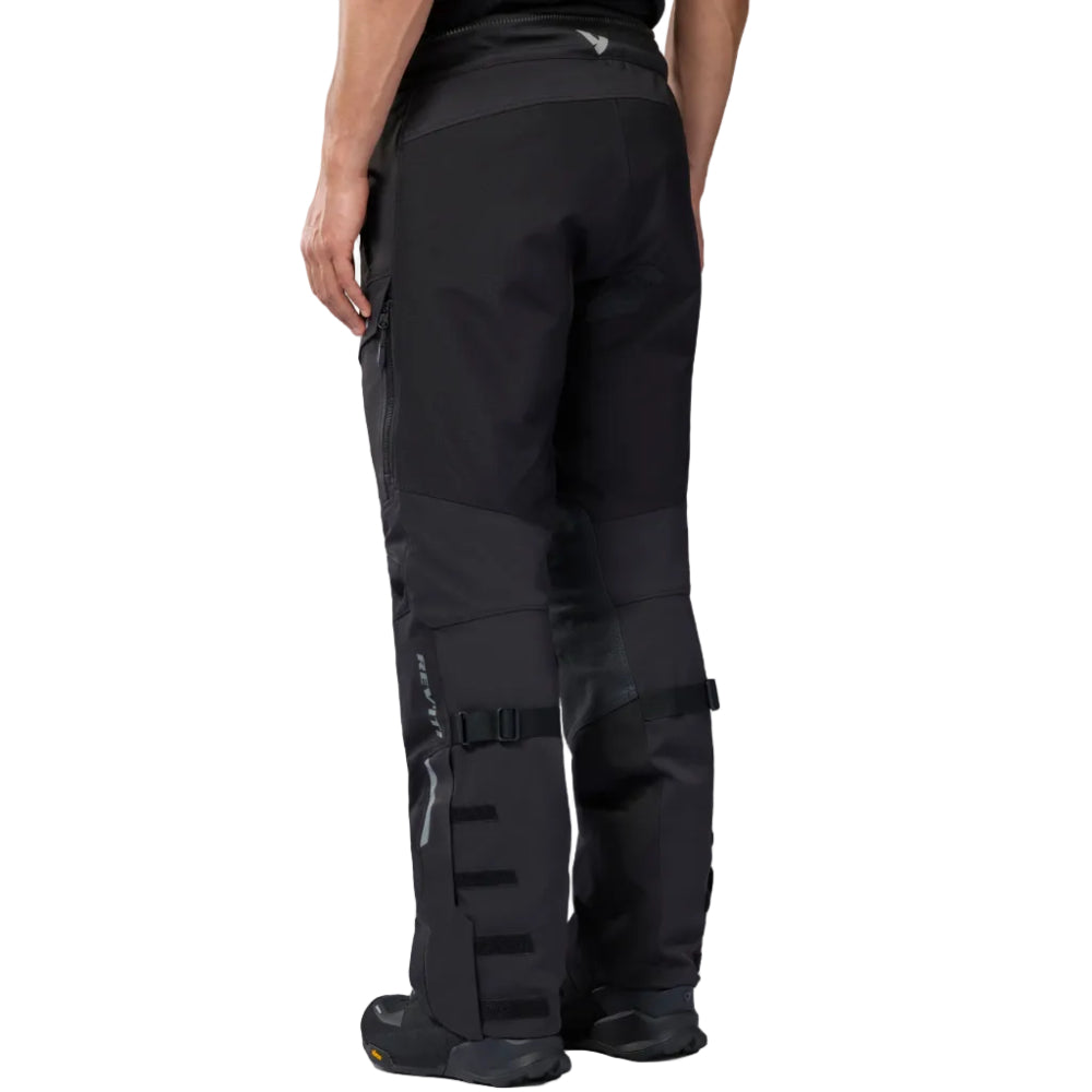 Rear view of a person wearing black technical pants with integrated boot covers and straps, shown against a plain white studio background.