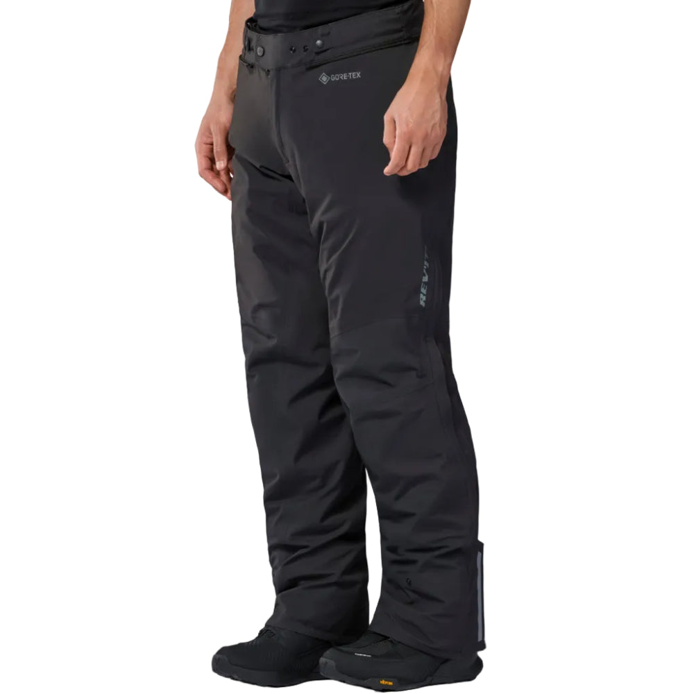 A person wearing black Gore-Tex snow pants with integrated boot covers, shown from the waist down against a clean white studio background.