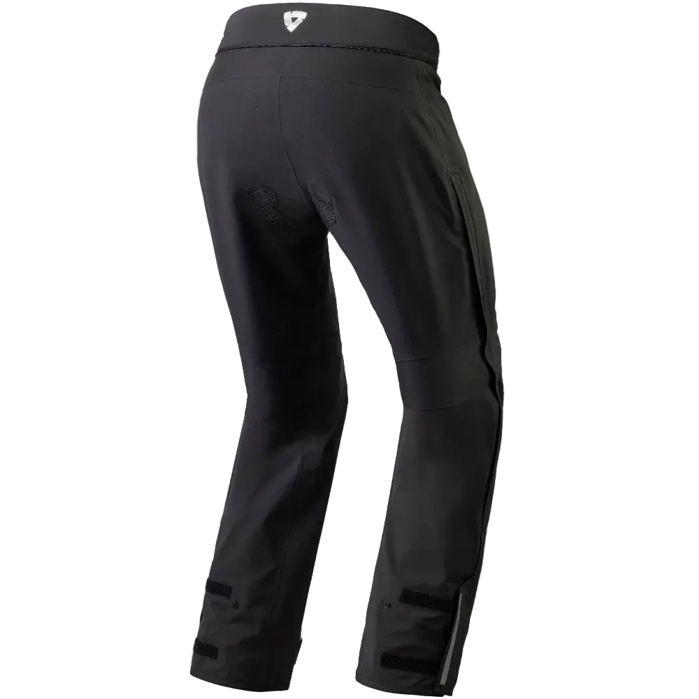 Black technical pants shown from the rear three-quarter view, featuring a high waistband with a small white shield logo and reinforced side paneling, on a solid white background.