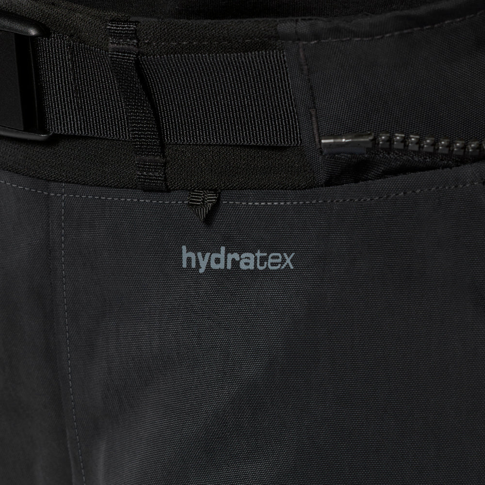 Close-up of black technical pants showing the waistband with a black belt, a zipper pocket, and the 'hydratex' logo printed in light gray on the front thigh.