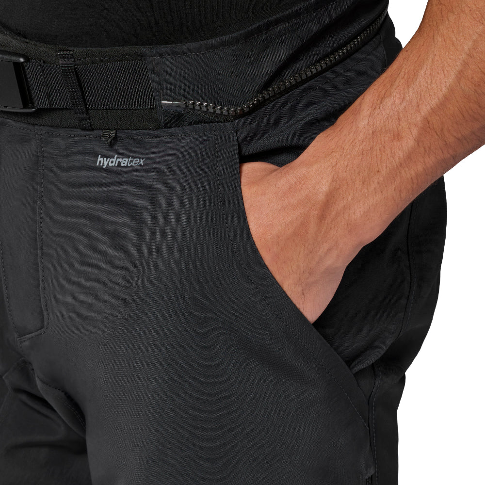 Close-up of a person wearing black pants with a side zipper, belt, and a small white 'hydrotex' logo near the pocket, with one hand in the pocket against a white background.