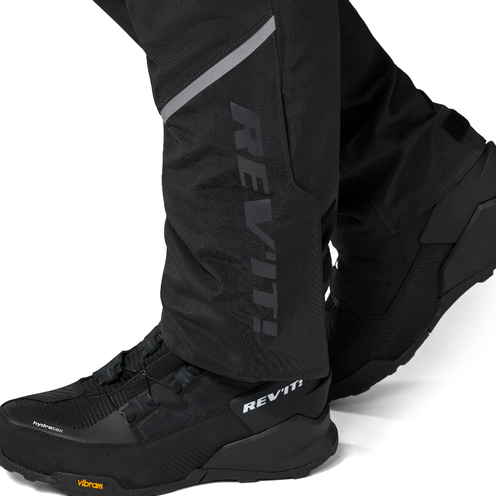 Black REV'IT! outdoor pants shown on a person's lower legs, paired with black shoes, against a clean white background.