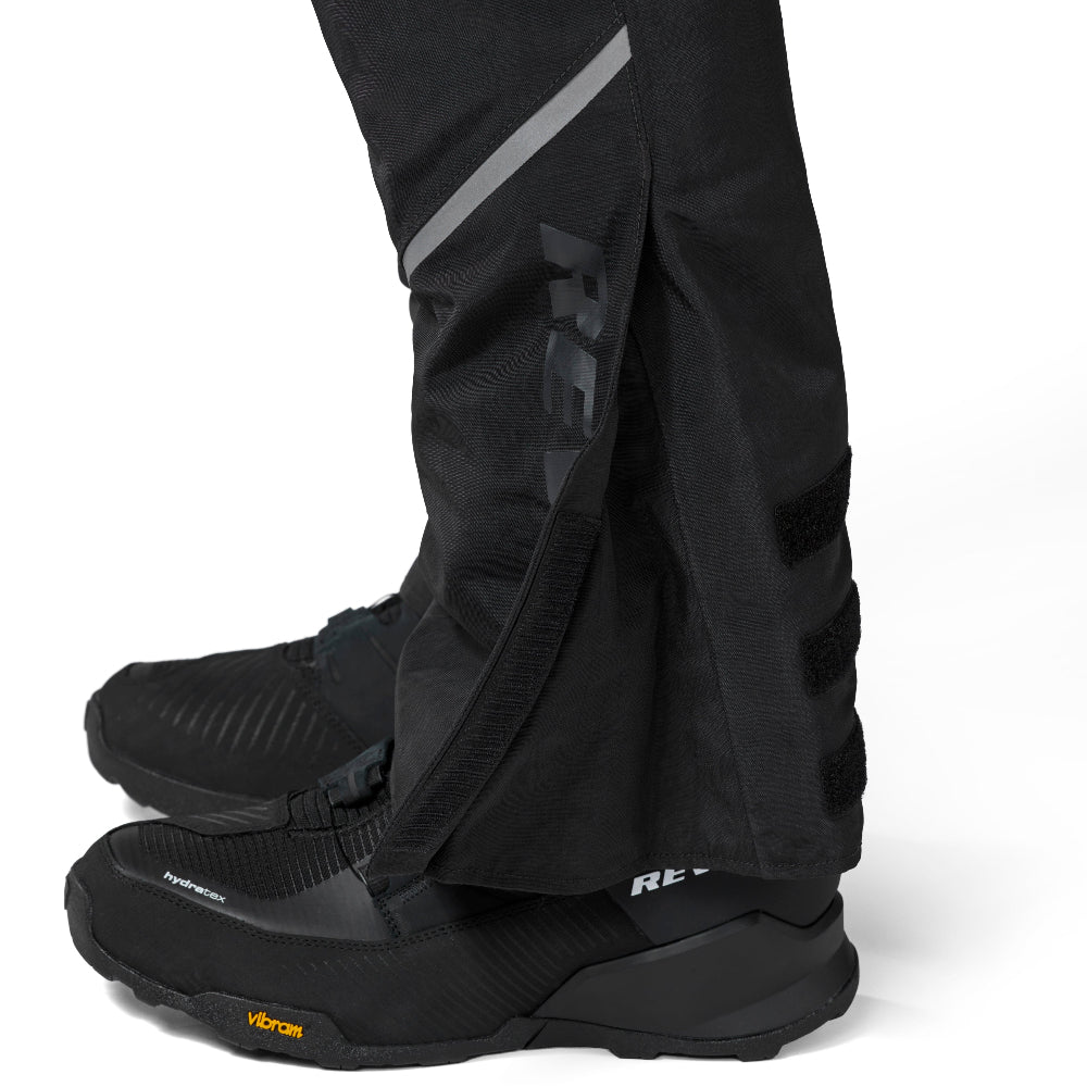 Black technical pants with reflective gray diagonal stripes and a large dark logo on the lower leg, shown worn with black boots against a clean white background.