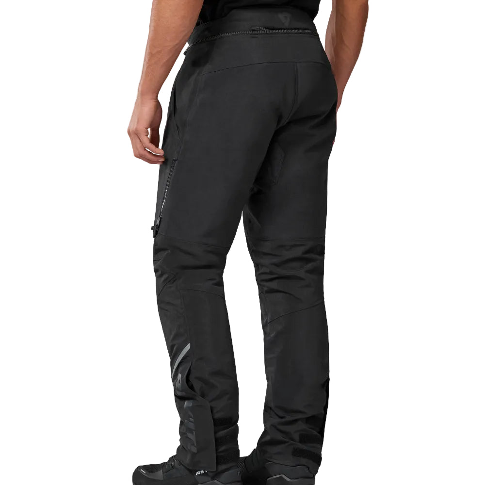 Rear view of a person wearing black technical outdoor pants and black boots against a plain light gray studio background.