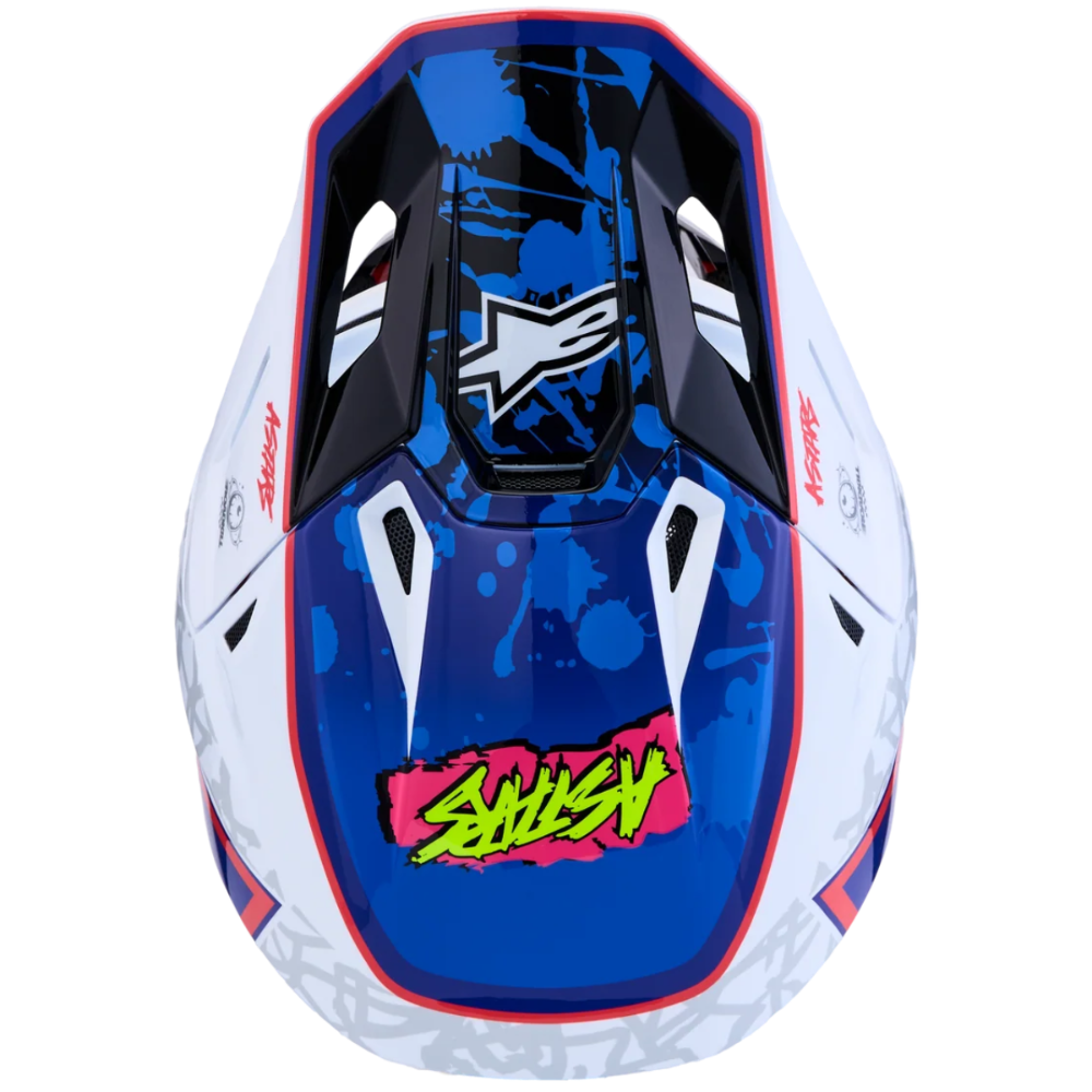 A motocross-style full-face helmet shown from the front on a plain white background. The helmet has a glossy blue visor with black splatter graphics, a white lower section, red trim, and a bright pink and yellow graphic near the bottom center.
