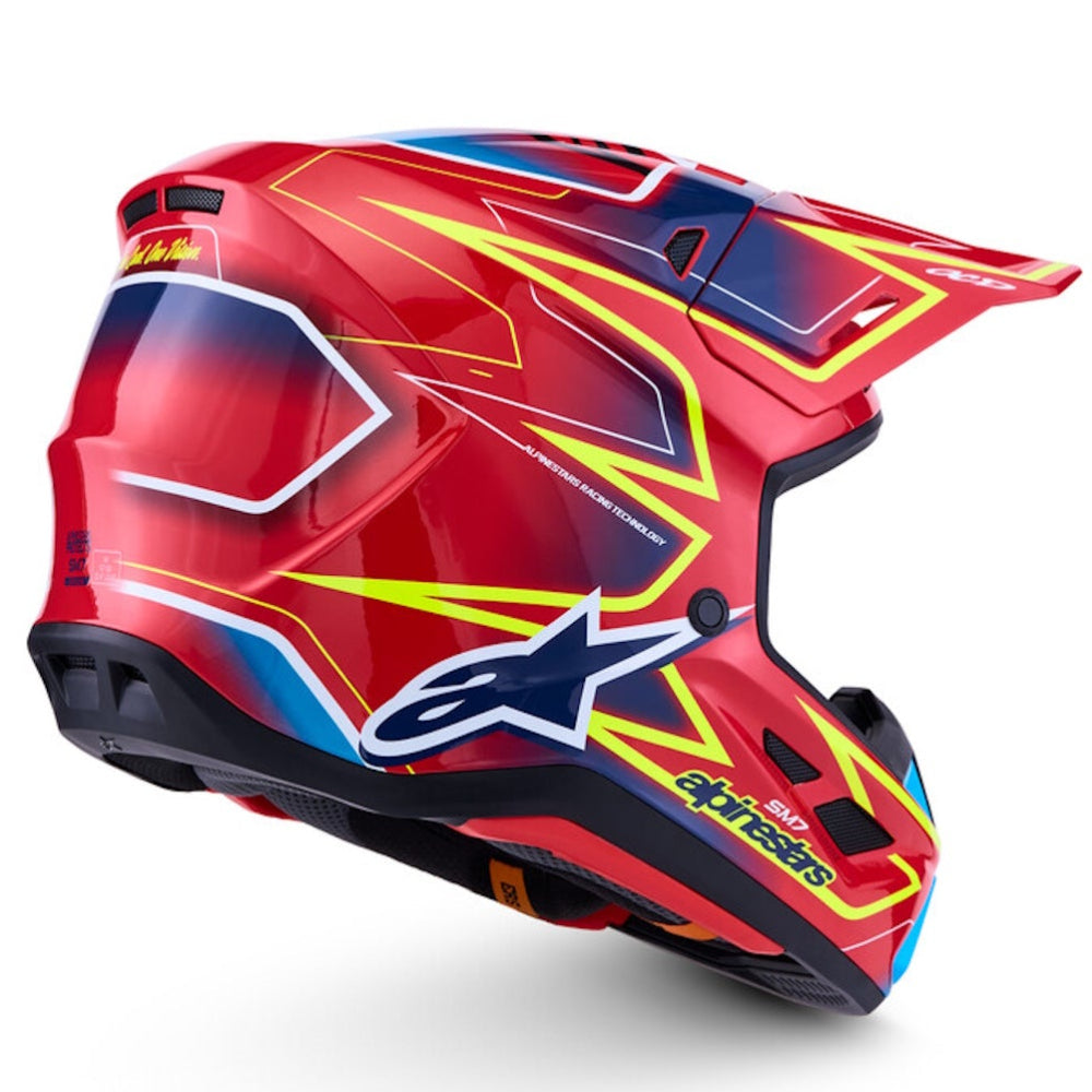A red Alpinestars motocross helmet with yellow, blue, and white graphic accents, shown from a three-quarters angle on a white background.