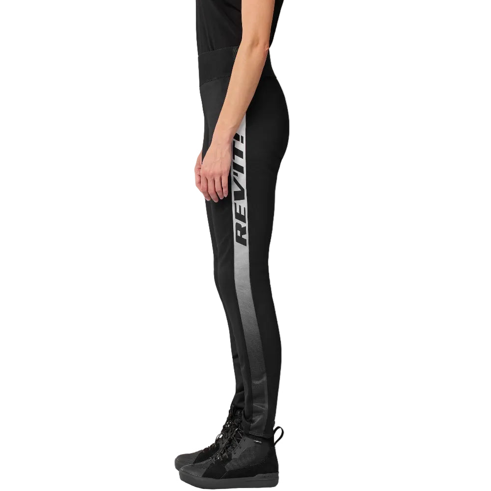 Side view of a person wearing black leggings with a vertical gray and white REV'IT! logo stripe down the left leg, paired with black high-top sneakers, against a light gray studio background.