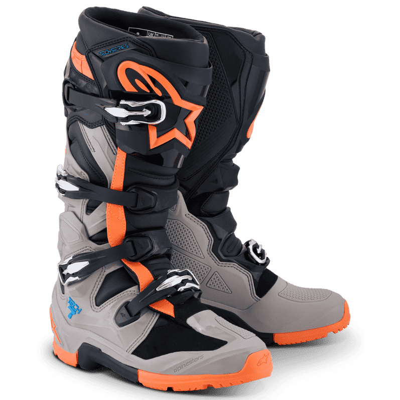 A pair of Alpinestars motocross boots in a Black/Warm Grey/Orange Fluo colorway, featuring the signature white Alpinestars star logo on the side, set against a clean white background.